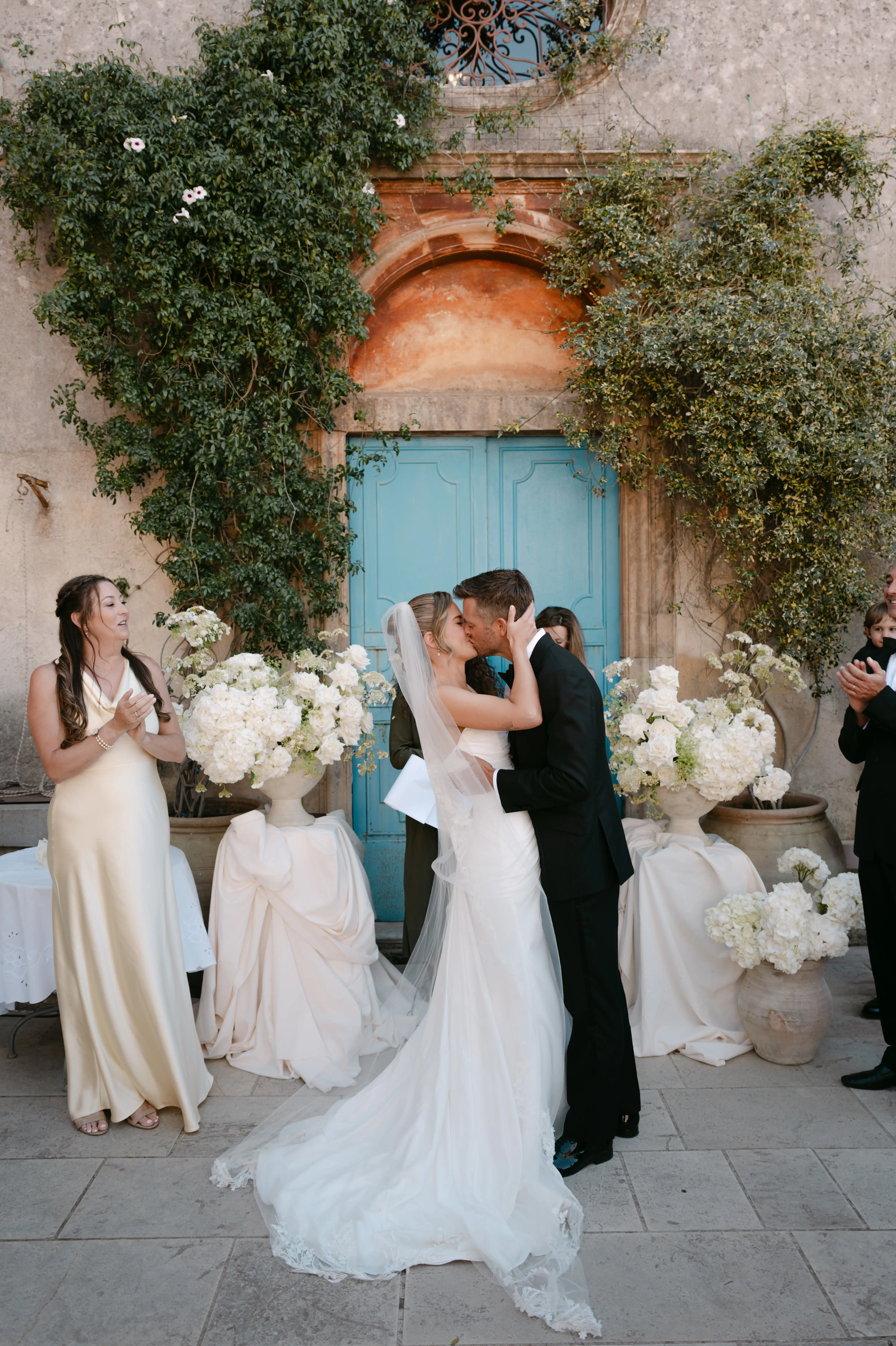 Bride and groom sharing their first kiss surrounded by white floral arrangements at a luxury Italian wedding venue.